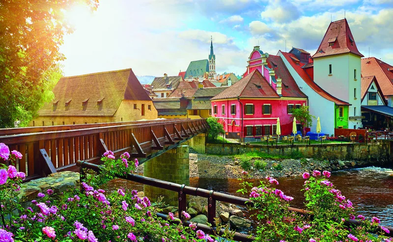 Czech Krumlov, (Cesky Krumlov), Czech Republic. Wooden bridge over river Vltava. Vintage picturesque old town with colorful houses and chapel of church. Rose fl