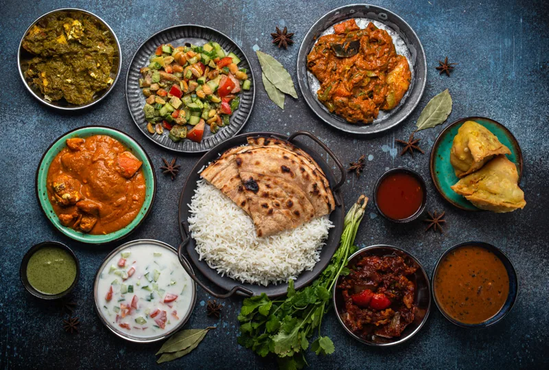 Assorted Indian ethnic food buffet on rustic concrete table from above: curry, fried samosa, rice biryani, dal, paneer, chapatti, naan, chicken tikka masala, tr