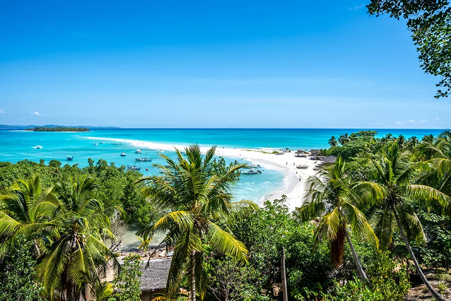 Description: White paradise sand beach with tourists leading into the turquoise blue, clear sea with boats photographed from hill on green palm tree island. Nos