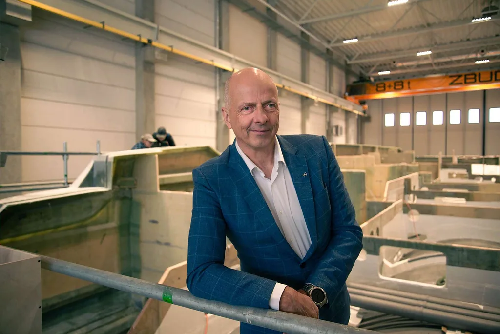 A man in a blue suit is standing in a Sunreef Yachts shipyard. The background shows yacht parts and two people working.