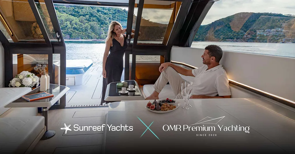 A couple enjoying time on a luxurious Sunreef Yachts vessel. The interior is elegant, featuring a table with snacks and drinks. Large windows provide a view of