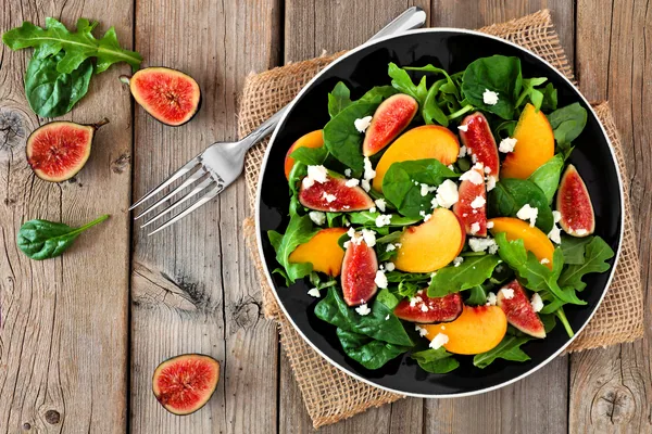 Autumn salad of arugula, spinach, figs and goat cheese in a black plate, overhead scene on a rustic wooden background